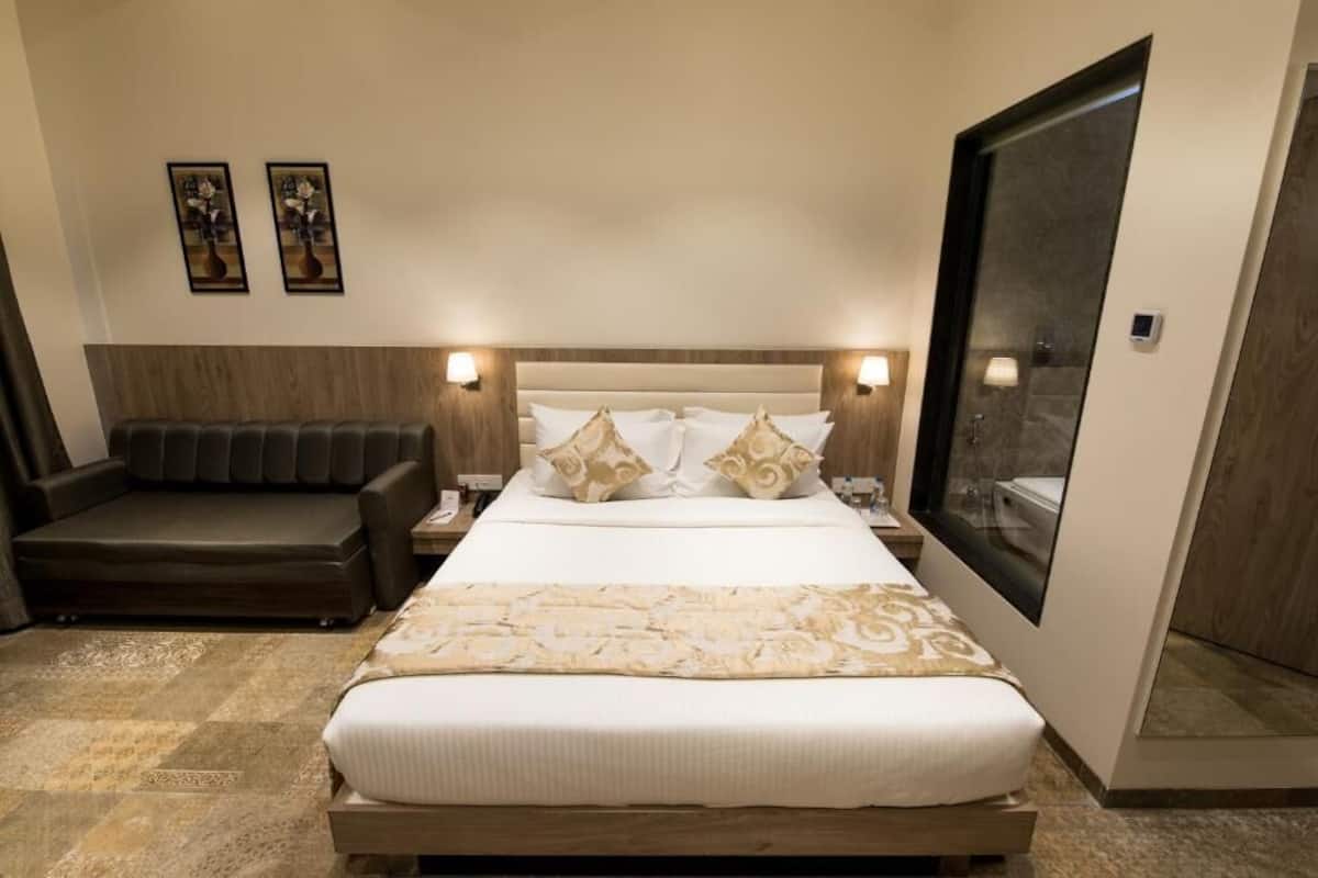 Quarto de hotel com uma cama grande, um sofá, duas luminárias fixadas na parede e uma janela ampla.