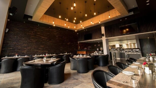 Restaurante moderno com pé-direito alto, vigas aparentes e luminárias pendentes. Mesas dispostas com toalhas brancas e cadeiras pretas.