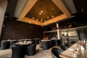 Un restaurante moderno con techos altos, vigas a la vista e iluminación colgante. Las mesas están dispuestas con manteles blancos y sillas negras.