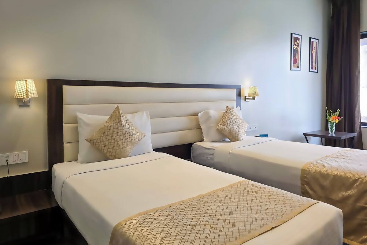 Quarto de hotel com duas camas, cada uma com cabeceira, roupa de cama branca e almofadas decorativas. Há luminárias na parede e uma pequena mesa com um vaso de flores.