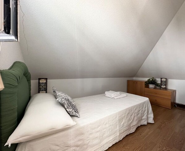 A small, neatly arranged bedroom with a single bed, a green couch, a wooden dresser, and a small table with a plant.