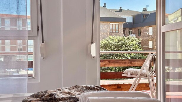 A bedroom with a bed, a window, and a view of a balcony with a chair and a wooden railing. - El Patuco - 30013 (Somo)