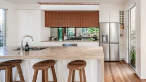 A modern kitchen with a central island, bar stools, a stainless steel refrigerator, and a wooden cabinet above the sink. - Headlands Hideaway, Moffat Beach Dog Friendly Home (Caloundra Region)