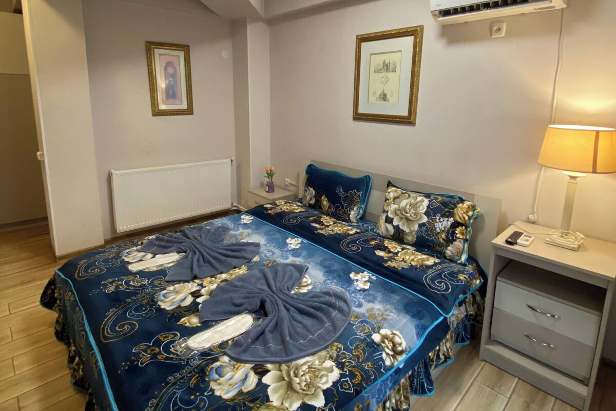 A bedroom with a large bed, a bedside table with a lamp, an air conditioning unit, and framed pictures on the wall.