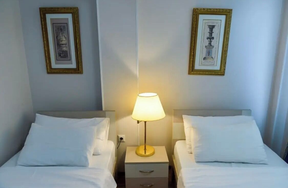 A hotel room with two single beds, a bedside table with a lamp, and two framed pictures on the wall.