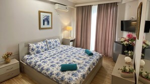 A bedroom with a bed, bedside table, lamp, air conditioning unit, curtains, and a TV. - Grand Central (Tsqaltubo)