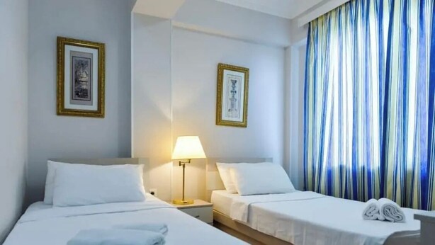 A hotel room with two beds, a nightstand with a lamp, and a window with striped curtains. - Grand Central (Tsqaltubo)