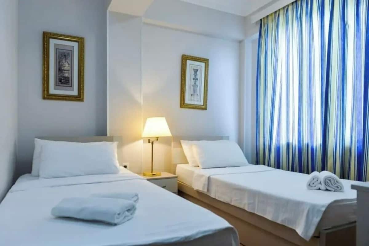 A hotel room with two beds, a nightstand with a lamp, and a window with striped curtains.