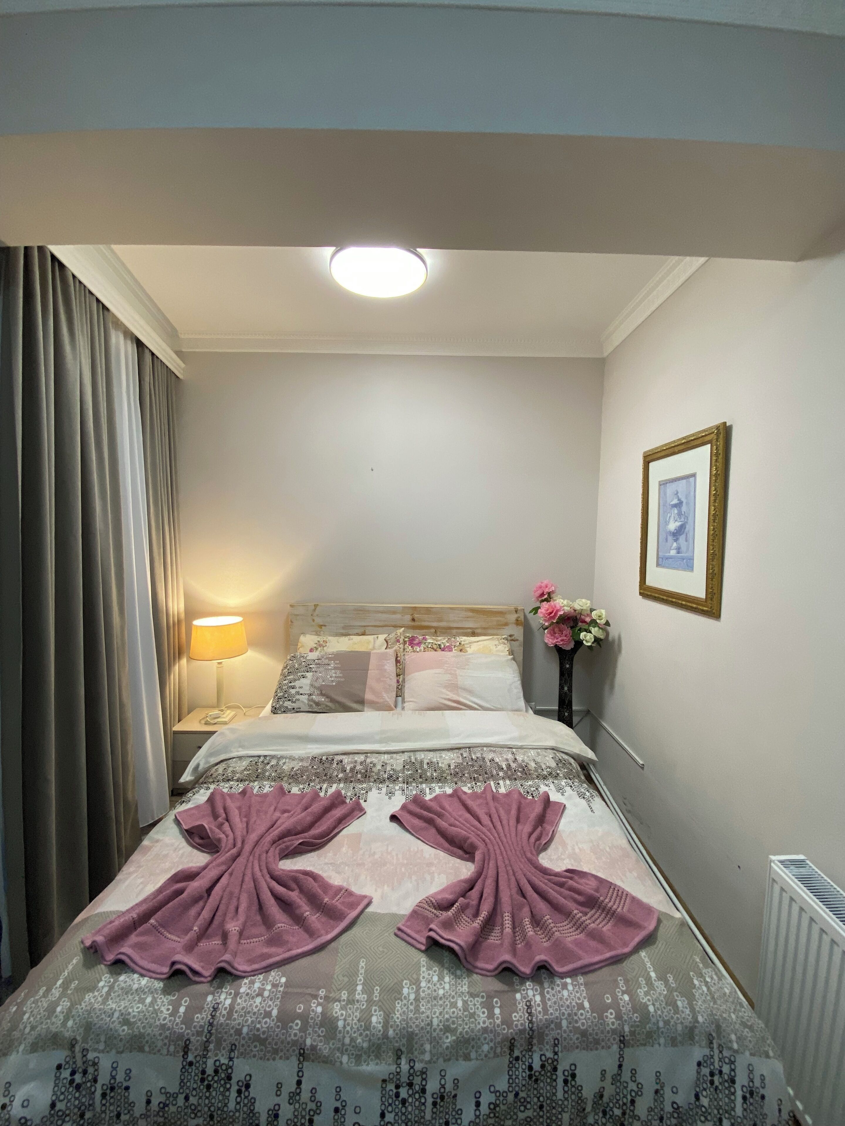 A hotel room with a bed, a nightstand with a lamp, a framed picture on the wall, and a window with curtains.