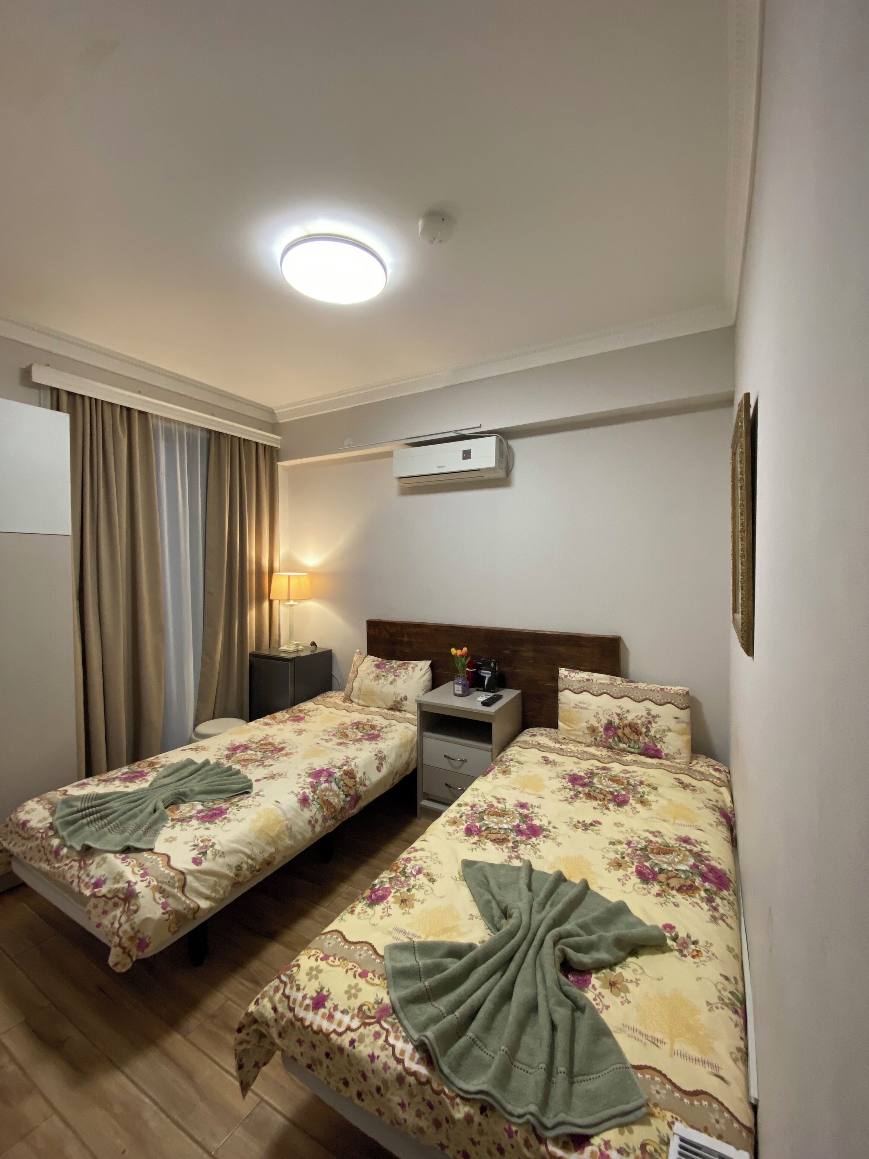 Habitación de hotel con dos camas individuales, una mesita de noche con lámpara, un equipo de aire acondicionado y una ventana con cortinas.