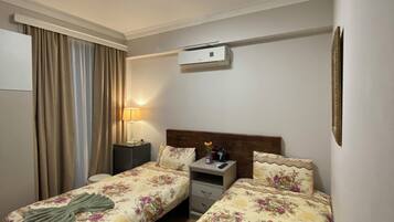 Habitación de hotel con dos camas individuales, una mesita de noche con lámpara, un equipo de aire acondicionado y una ventana con cortinas.