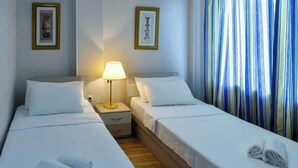 A hotel room with two beds, a nightstand with a lamp, and a window with striped curtains. - Grand Central (Tsqaltubo)