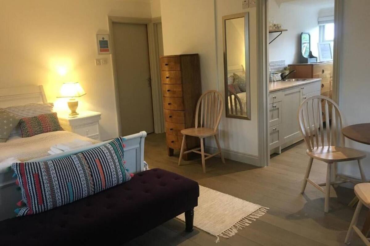 Et soveværelse med en seng, en sengebordslampe og en mørk bænk. Der er en træstol og et spejl på væggen. Et køkken er synligt gennem en åben dør.