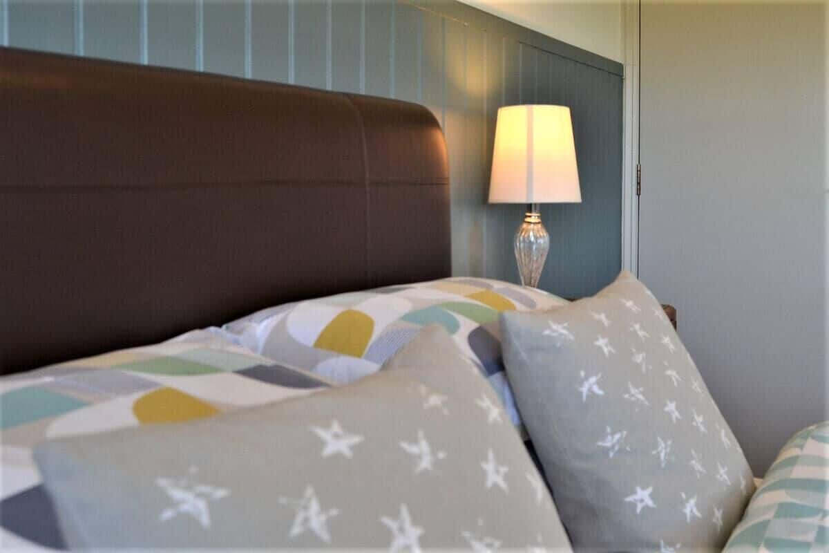 En pænt redt seng med mønstrede puder og en sengegavl, en sengetlampe, og en væg med lodrette linjer.