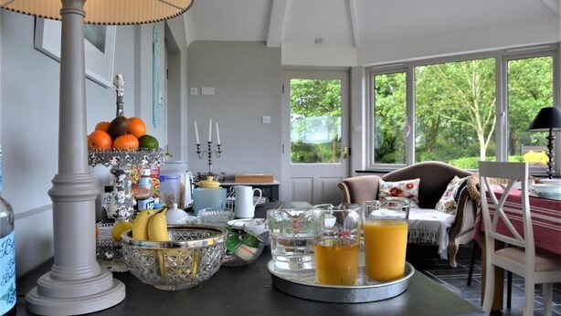 A breakfast setup with fruits, drinks, and a table in a bright room with large windows.