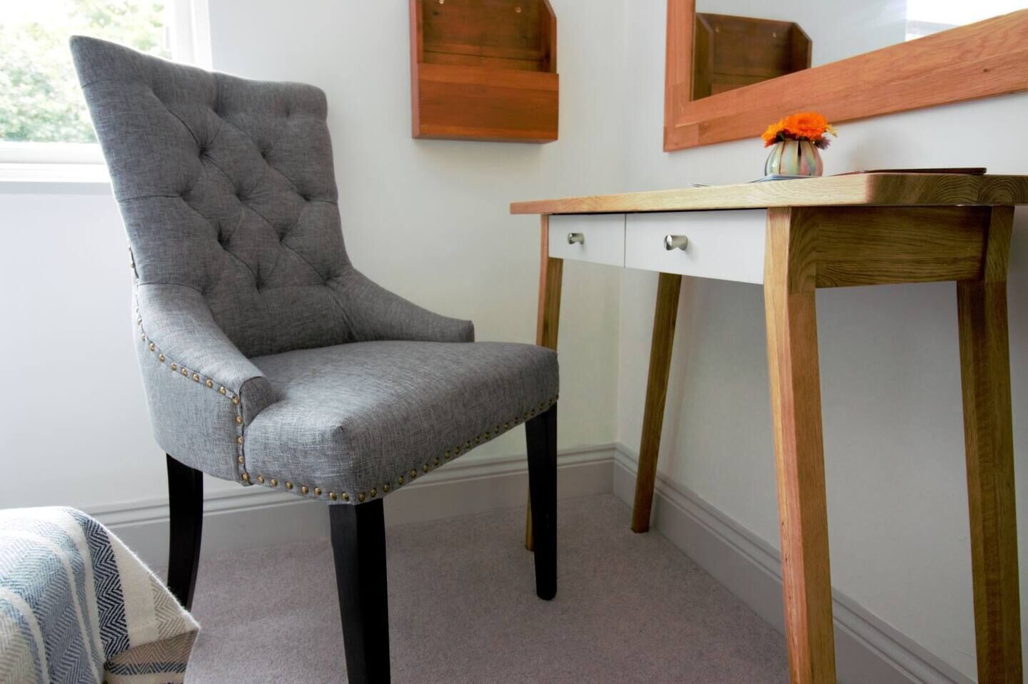 A grey tufted chair with metal studs, a wooden desk with drawers, and a mirror above it.