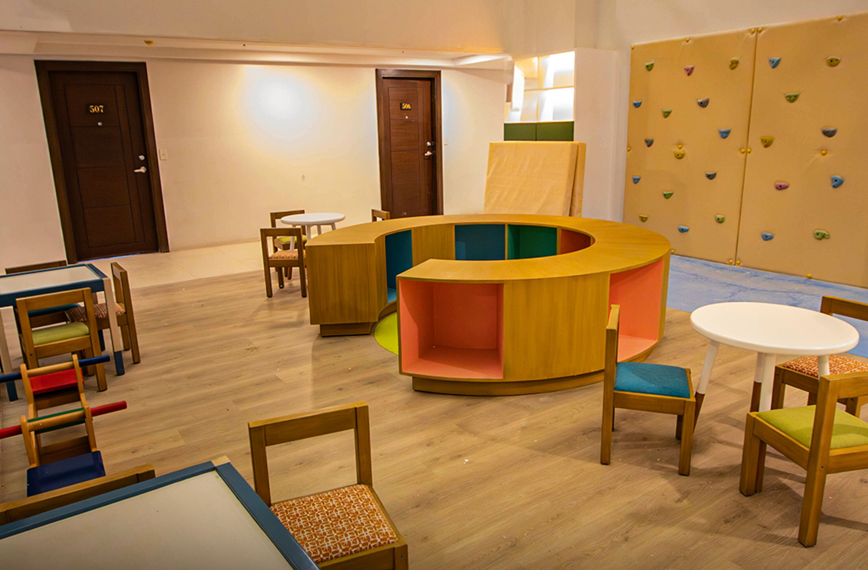 A children's playroom with a circular seating area, wooden tables, and climbing wall.