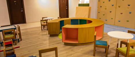 A children's playroom with a circular seating area, wooden tables, and climbing wall.