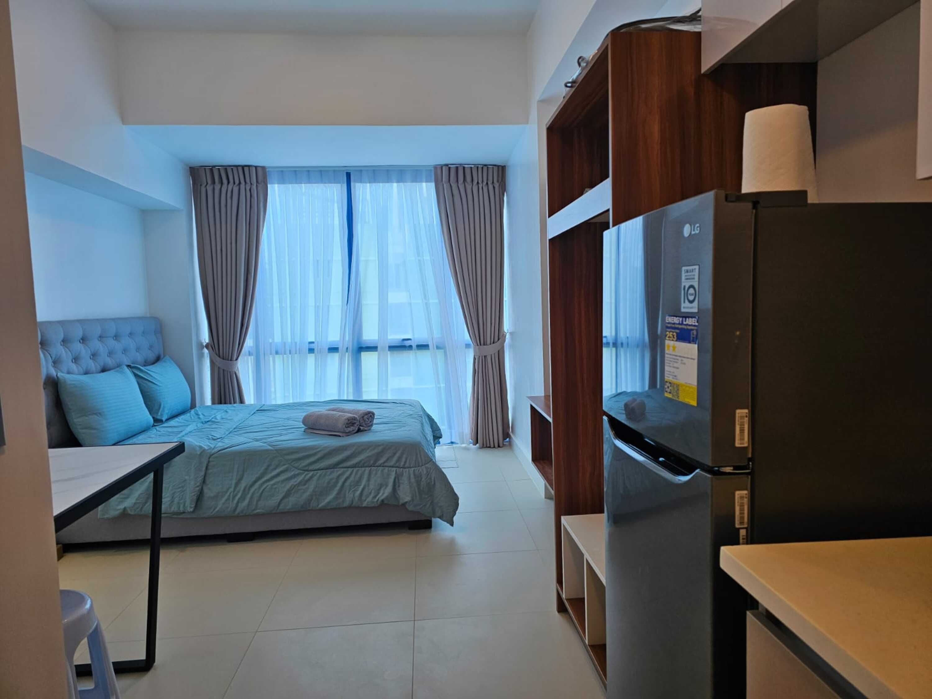 A hotel room with a bed, a small table, a refrigerator, and a view of the outside through sheer curtains.