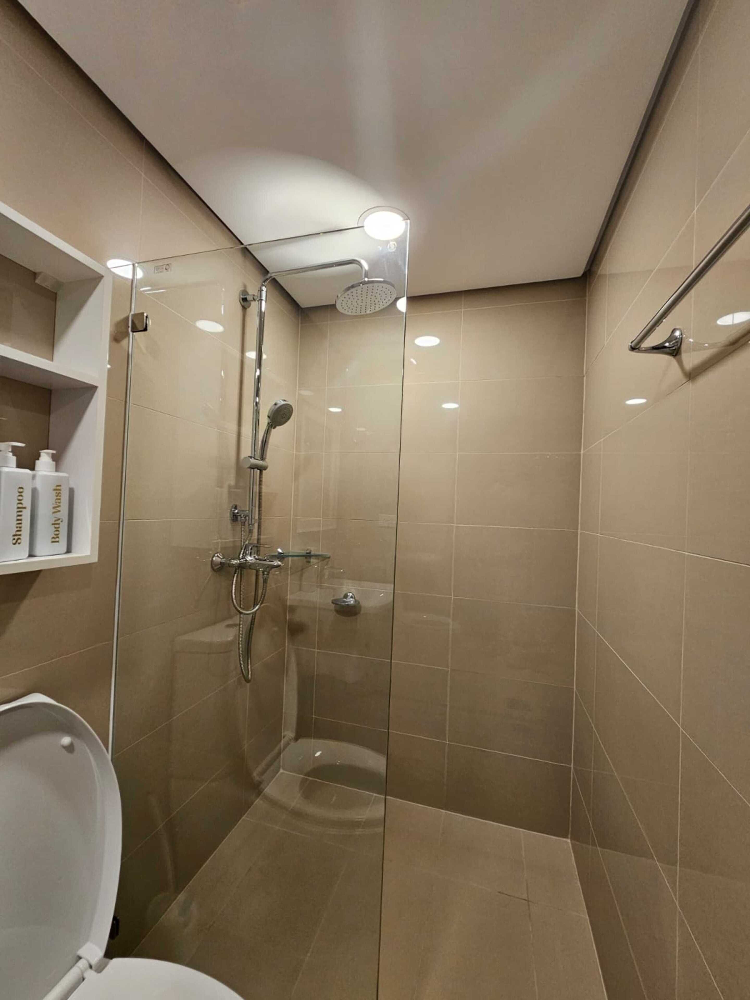 Egy modern fürőszoba üvegfalas zuhanyzóval, WC-vel és egy polccal, amelyen a fürdőszobai kellékek találhatók.