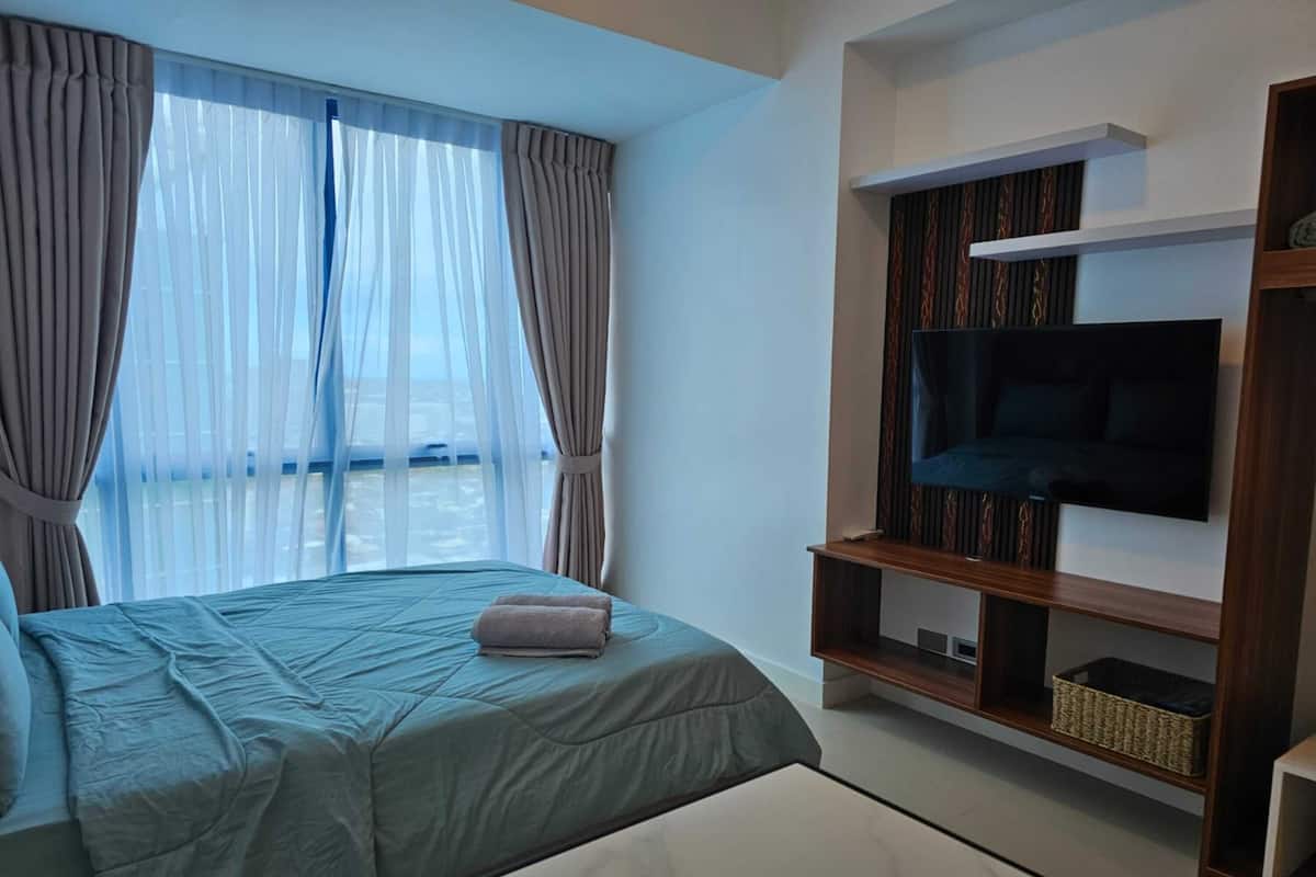 Un dormitorio con una cama, un televisor montado en un estante de madera y vista al mar a través de cortinas translúcidas.