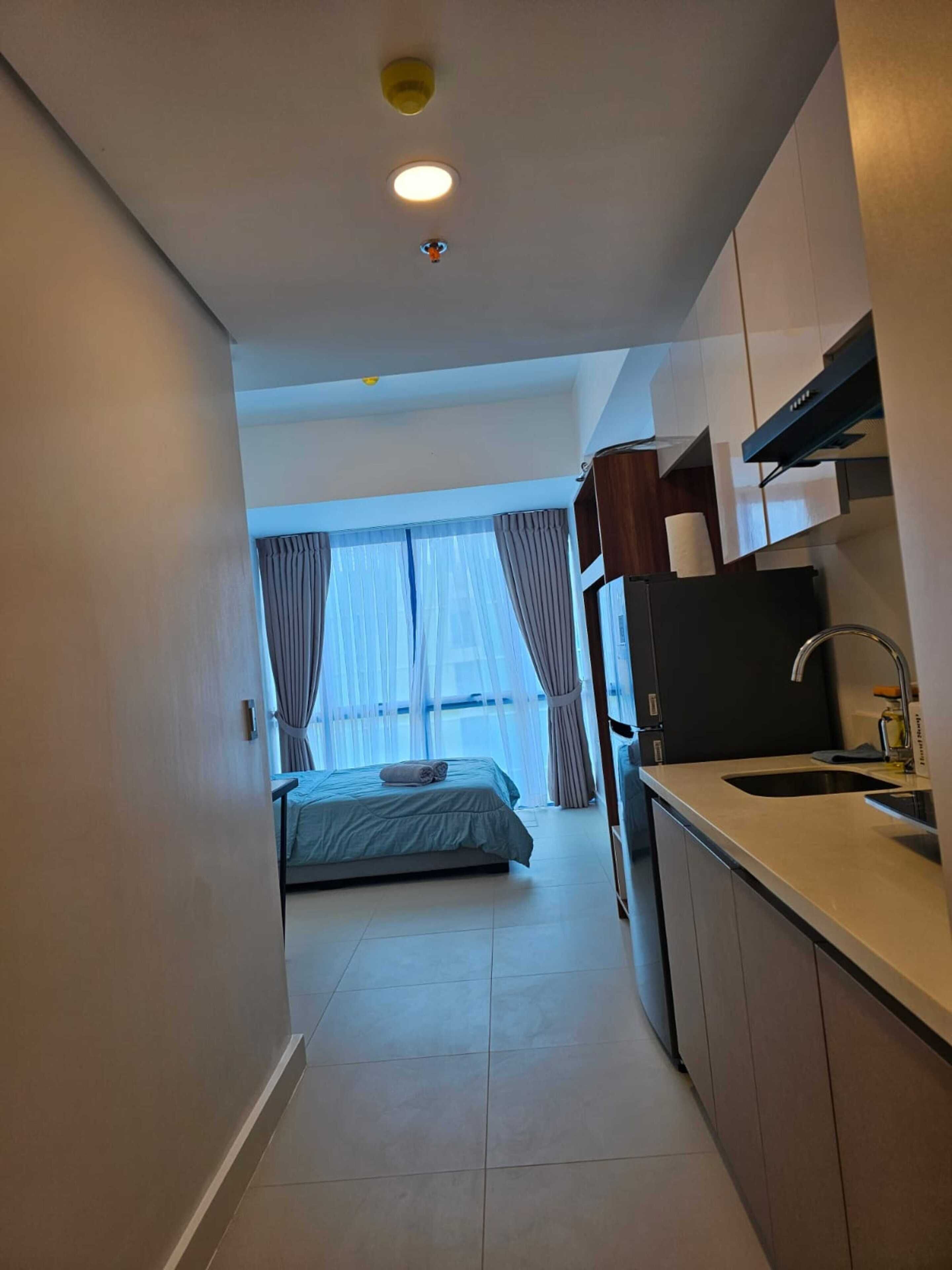 A narrow hallway leading to a bedroom with a bed and a bathroom. The room has a kitchenette with a sink and cabinets.