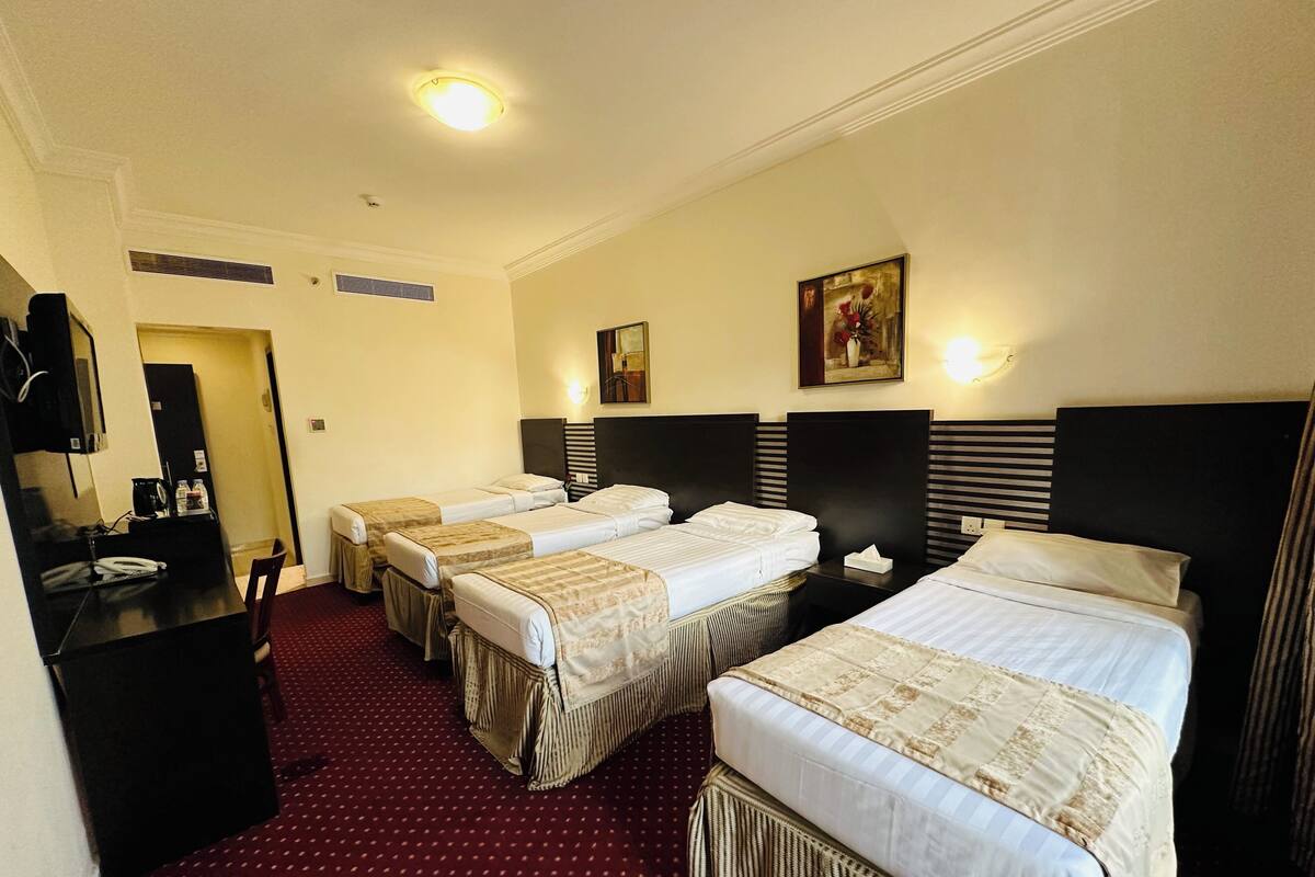 Habitación de hotel con tres camas, un escritorio, una silla, un radiador, un cuadro y una lámpara de techo.