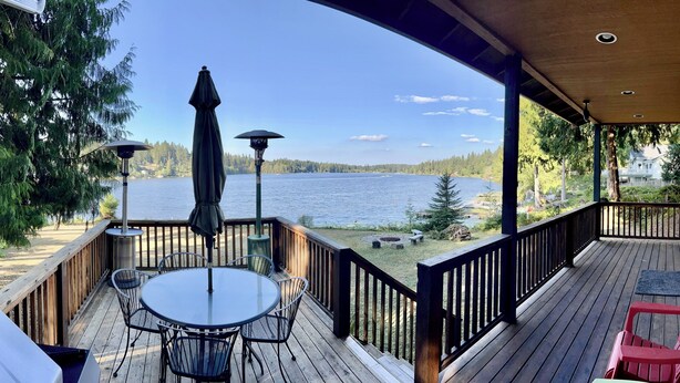 A wooden deck with a table and chairs, overlooking a lake with trees and a distant house.