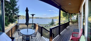 A wooden deck with a table and chairs, overlooking a lake with trees and a distant house. - Serenity Shores: Lakefront Vacation Home with Indoor Playhouse! (Eatonville)