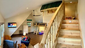 A loft-style room with a wooden staircase, a television, a desk, and a window with blinds.