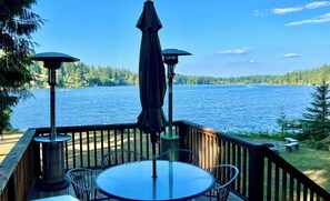 A wooden deck with a round table and chairs, two umbrellas, and a view of a lake. - Serenity Shores: Lakefront Vacation Home with Indoor Playhouse! (Eatonville)