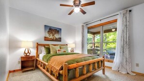 A bedroom with a wooden bed, two bedside tables, a ceiling fan, a large window with curtains, and a painting on the wall.