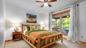 A bedroom with a wooden bed, two bedside tables, a ceiling fan, a large window with curtains, and a painting on the wall.