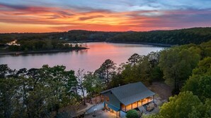 A lakeside cabin with a deck and a covered porch, surrounded by dense forest.