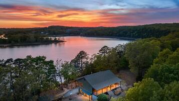 A lakeside cabin with a deck and a covered porch, surrounded by dense forest.