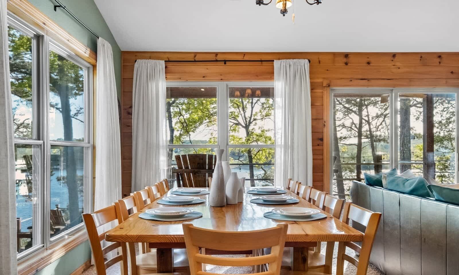 A dining area with a wooden table set for six, surrounded by chairs, with large windows offering a view of trees and a body of water.