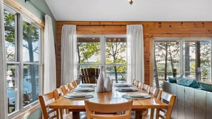 A dining area with a wooden table set for six, surrounded by chairs, with large windows offering a view of trees and a body of water.