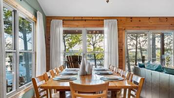 A dining area with a wooden table set for six, surrounded by chairs, with large windows offering a view of trees and a body of water.
