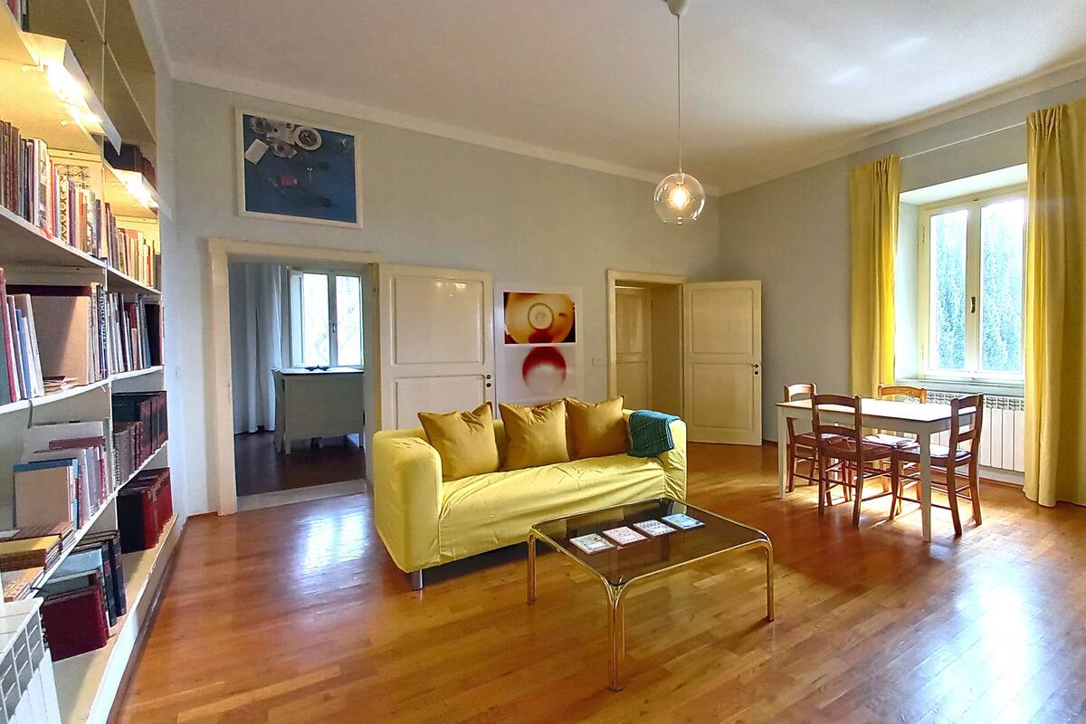 Una sala de estar con un sofá amarillo, una mesa de centro de vidrio, una mesa de comedor de madera con sillas, una estantería y un ventanal con cortinas.