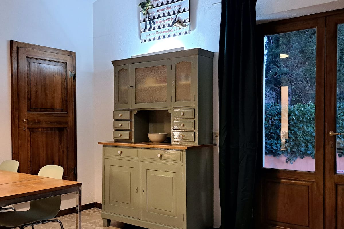 Una habitación con una mesa de madera, dos sillas, un armario con cajones, un televisor en la pared y una ventana con vistas a la vegetación.