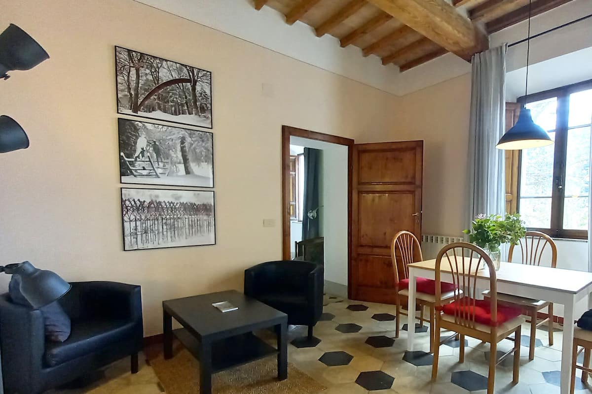 Una sala de estar acogedora con un sofá, una mesita y un comedor con sillas y una mesa. En la pared hay cuadros enmarcados, una lámpara y una ventana con cortinas.
