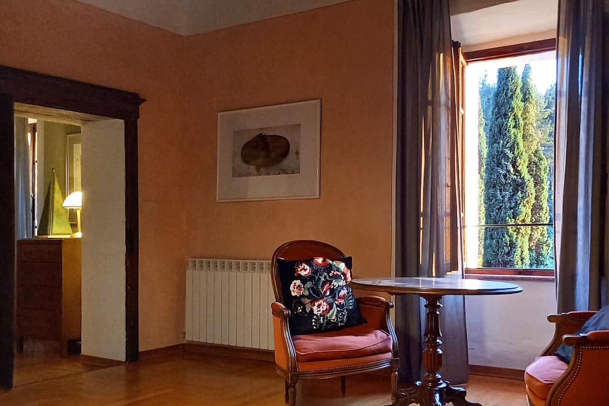 Una habitación con piso de madera, una mesa redonda, dos sillas con cojines estampados, un cuadro en la pared y una ventana con cortinas.