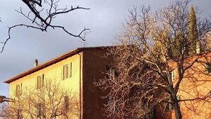 Ein zweistöckiges Gebäude mit gelber Fassade und rotem Dach. Vor dem Gebäude steht ein Baum und der Himmel ist wolkenlos.