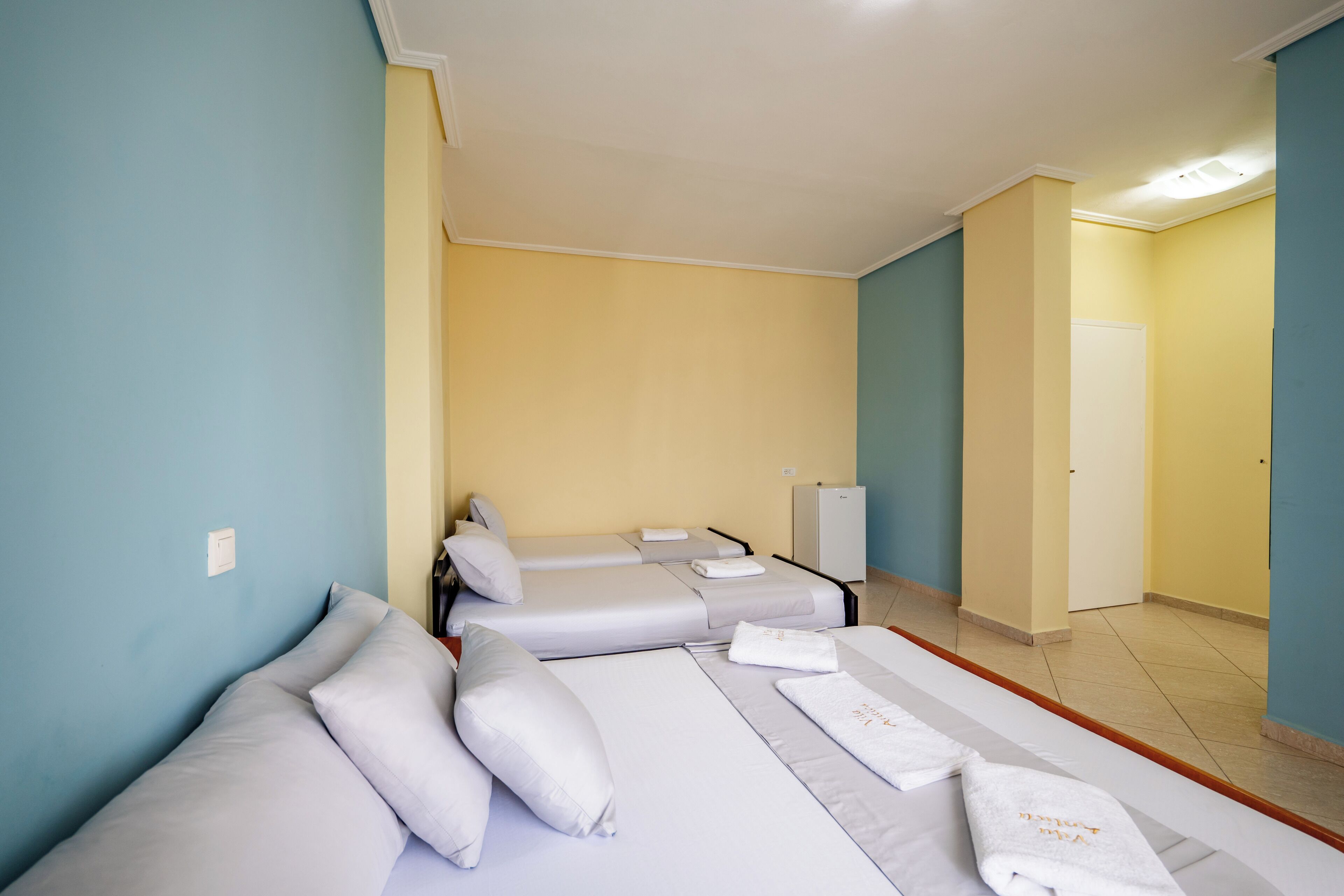 Habitación de hotel con tres camas, cada una con sábanas y almohadas blancas, un mueble pequeño y una puerta que da a otra habitación.