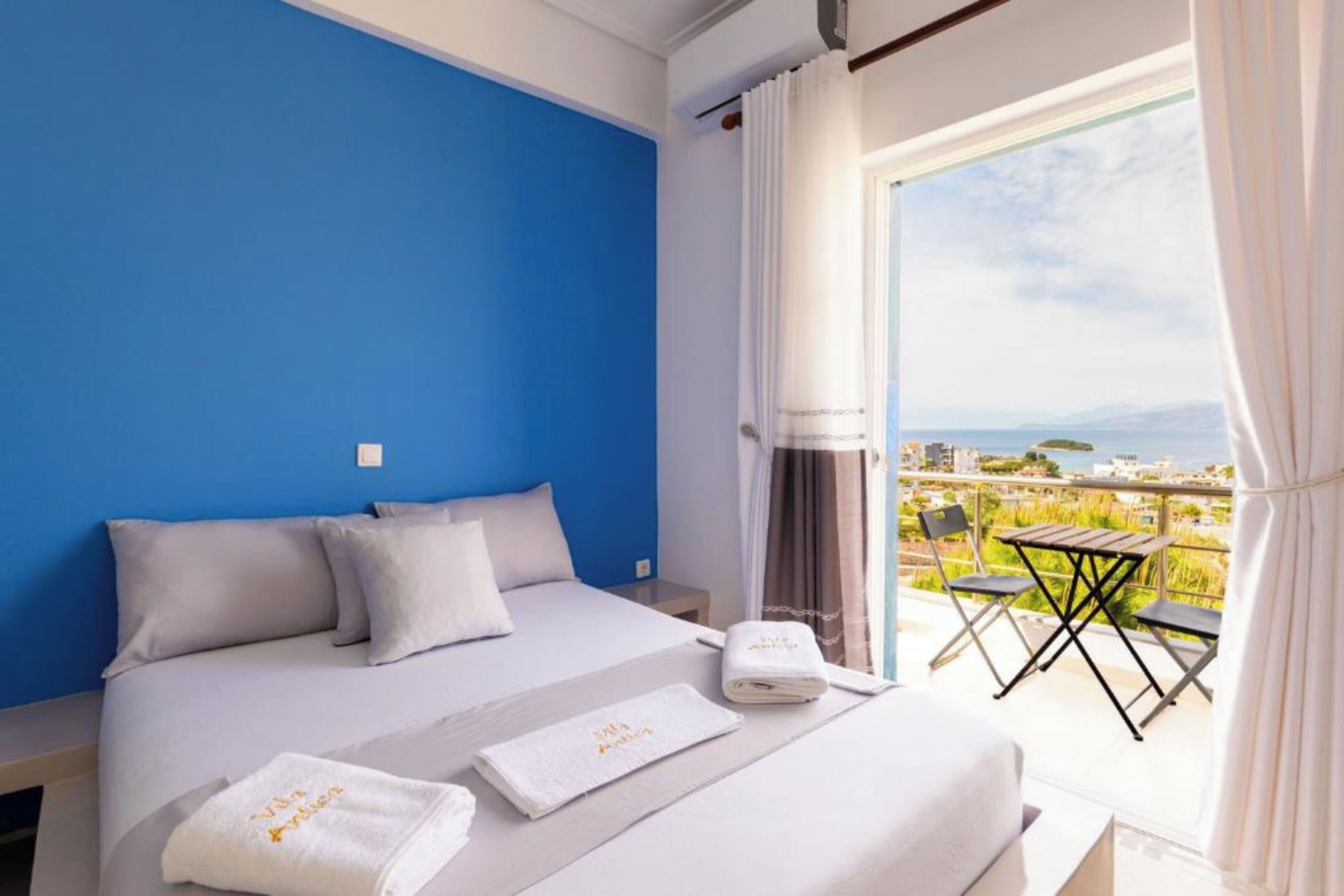 A hotel room with a blue accent wall, a bed with white linens, a balcony with a table and chairs, and a view of the sea and buildings.