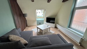 Ein gemütliches Wohnzimmer mit einer Couch, einem Couchtisch und einem Fernseher. Ein Fenster bietet Blick ins Freie.