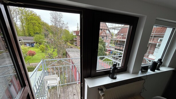 Ein Zimmer mit einem großen Fenster, das auf einen Garten und angrenzende Gebäude blickt. Es gibt einen Heizkörper, einen kleinen Tisch mit einer Lampe und einen Stuhl.