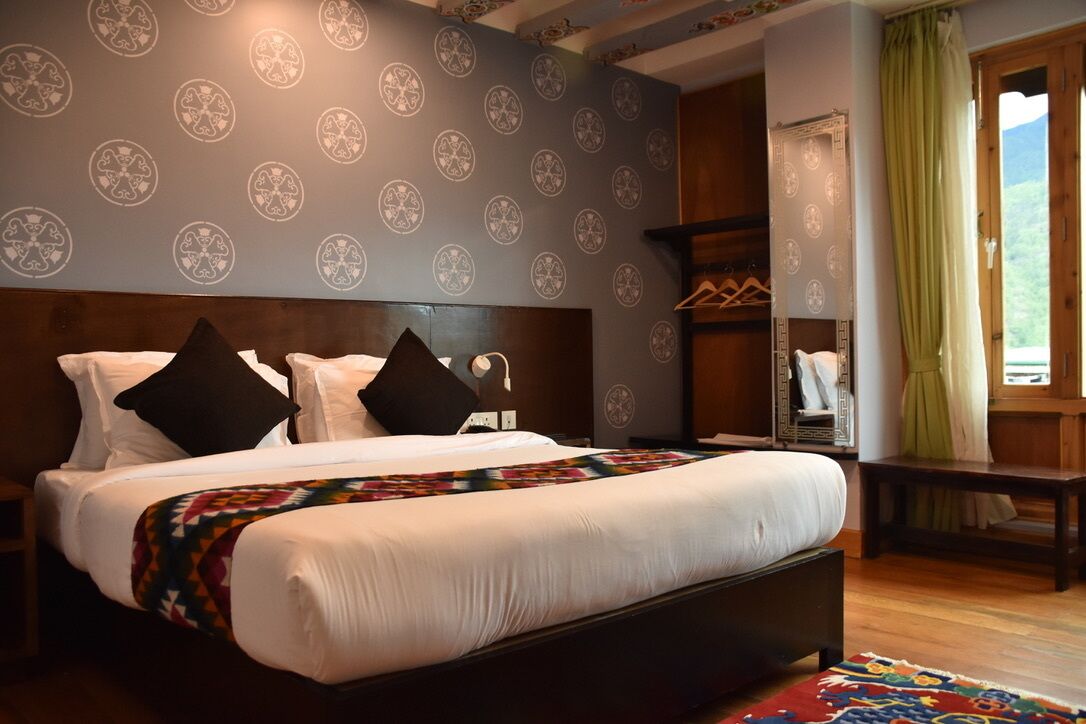 A bedroom with a large bed, a wooden headboard, a patterned wall, a mirror, and a view of the outdoors.