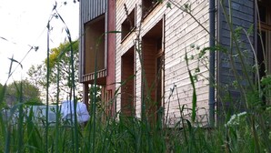 Een houten gebouw met twee verdiepingen, voorzien van een balkon op de bovenverdieping, omringd door hoog gras en begroeiing.