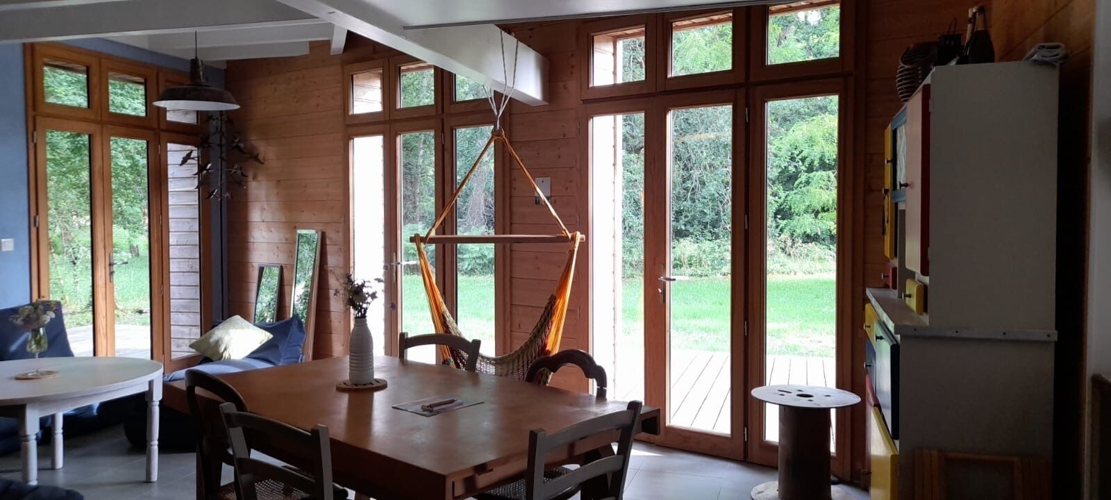A dining area with a wooden table and chairs, large windows, and a hanging light fixture.